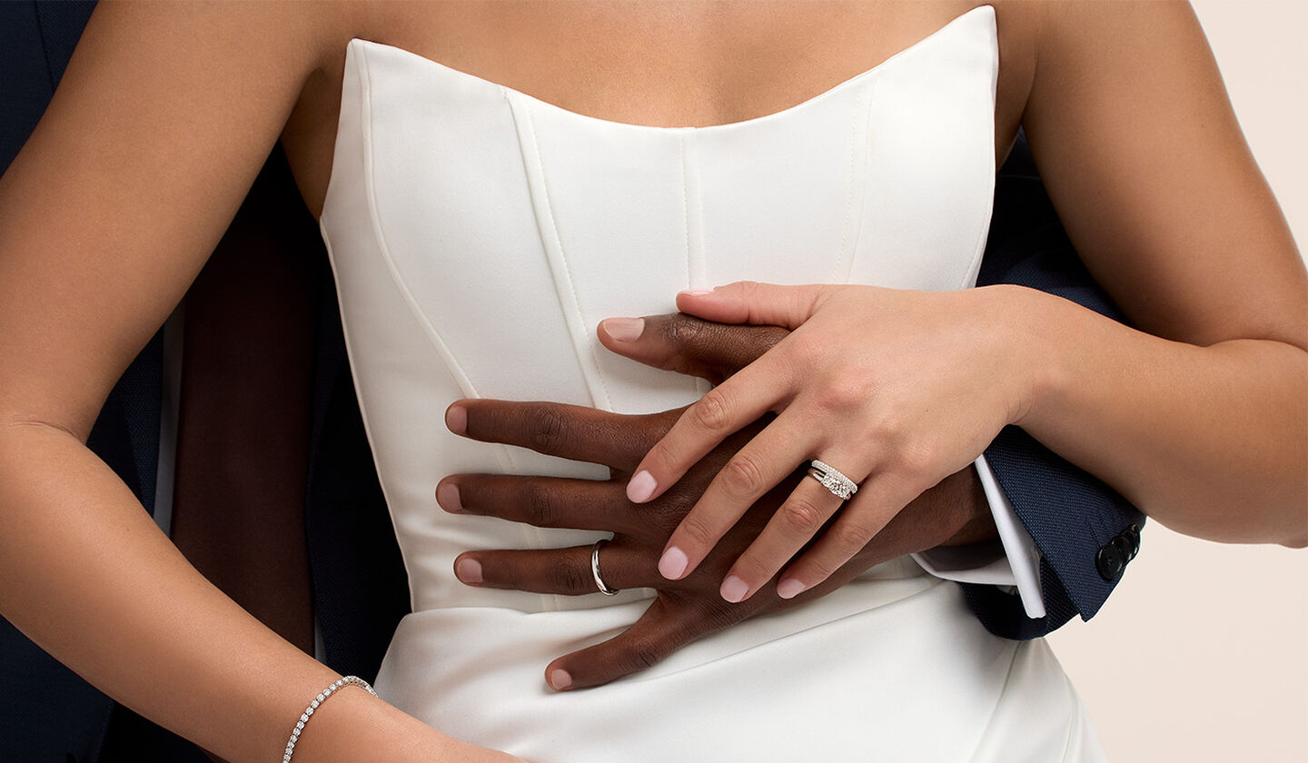 A close-up of a bride and groom embracing. The bride is wearing a strapless white wedding dress and a Birks diamond ring on her finger. The groom, dressed in a dark suit, wraps his arms around her waist. Both of their hands are gently intertwined, showcasing their Birks wedding rings.